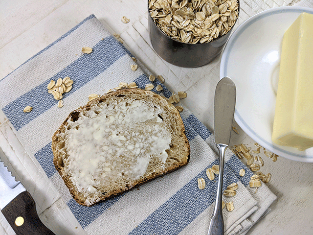 slice of maple oat sourdough sandwich bread on tea towel next to butter and cup of oats