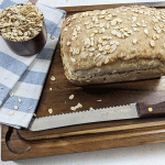 maple oat sourdough sandwich bread on cutting board with bread knife and cup of oats