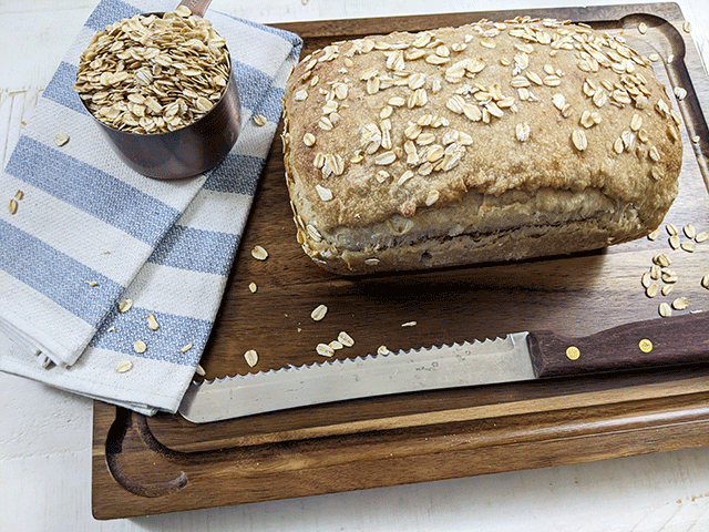 maple oat sourdough sandwich bread on cutting board with bread knife and cup of oats